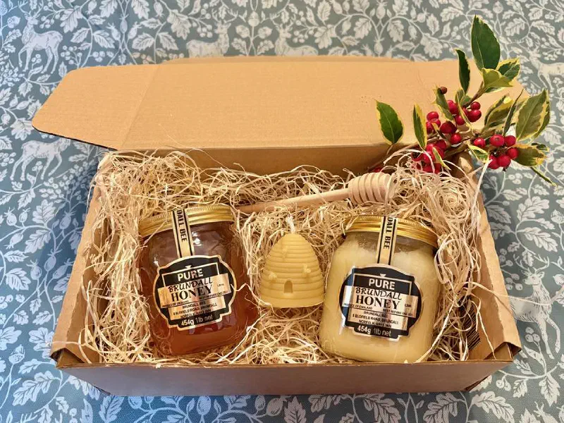 A gift box with jars of runny and soft set Brundall Bee honey, a beeswax skep candle and a wooden honey dibber, with a sprig of holly
