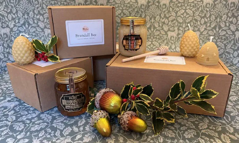 Brundall Bee gift boxes with jars of honey, beeswax pine cone and skep candles, a honey dibber, holly and gold acorns