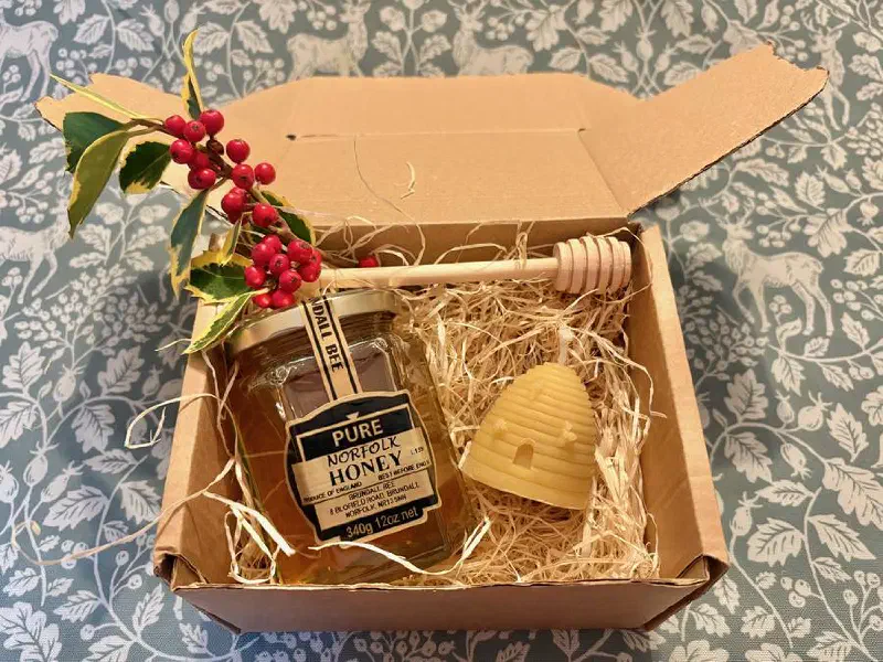 A gift box with a jar of Brundall Bee honey, a beeswax skep candle and a wooden honey dibber in wood wool, with a sprig of holly