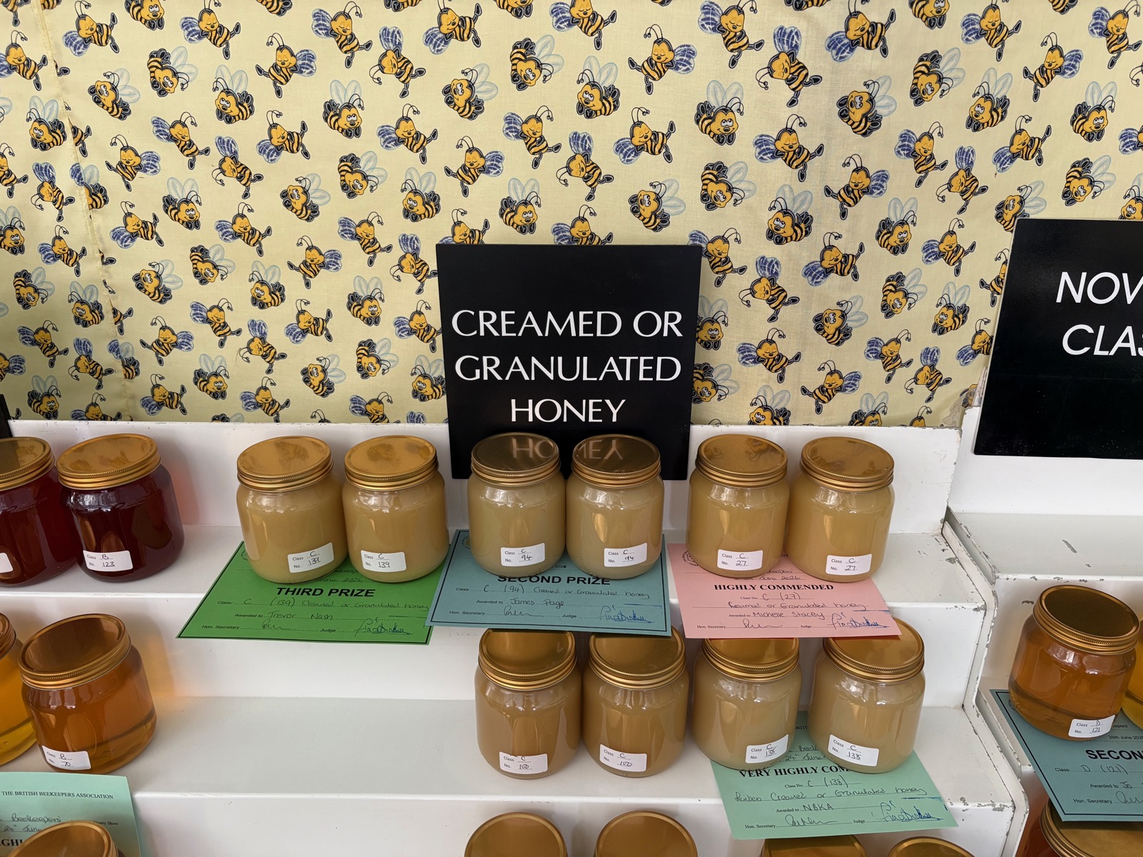 Honey show display shelves with jars of creamed honey and a second prize card visible, bee-print wallpaper behind